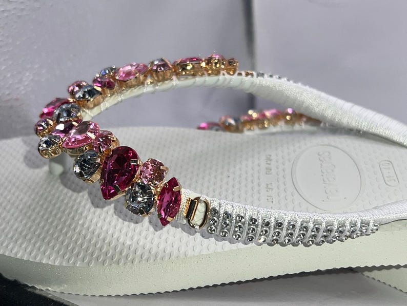 May include: A white flip-flop featuring a strap embellished with a variety of pink and dark gray faceted crystals in different shapes. The base of the strap includes a row of small, clear, round rhinestones set in a metallic frame.