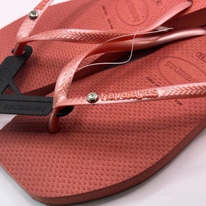K&ouml;nnte beinhalten: Nahaufnahme eines Paares korallenfarbener Havaianas-Flip-Flops. Die strukturierten Sohlen und Riemen sind in einem passenden Korallenton gehalten, mit dem Markennamen auf dem Riemen. Ein schwarzes Etikett ist an einem Flip-Flop befestigt.
