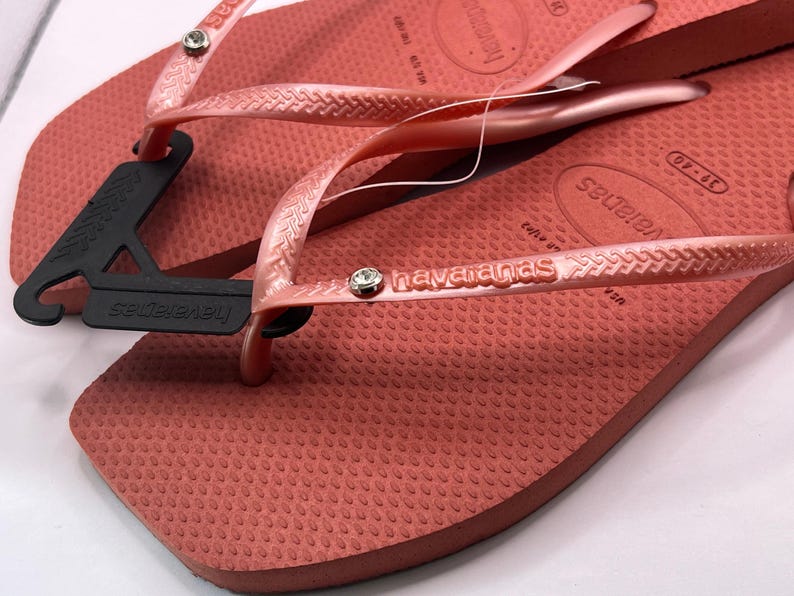 K&ouml;nnte beinhalten: Nahaufnahme eines Paares korallenfarbener Havaianas-Flip-Flops. Die strukturierten Sohlen und Riemen sind in einem passenden Korallenton gehalten, mit dem Markennamen auf dem Riemen. Ein schwarzes Etikett ist an einem Flip-Flop befestigt.