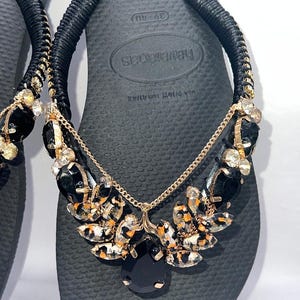 May include: Black flip-flops featuring black fabric-wrapped straps adorned with gold-tone chains and clusters of black and leopard-print faceted crystals. The footbed displays the brand name Havaianas and size 39-40, with a textured surface pattern.