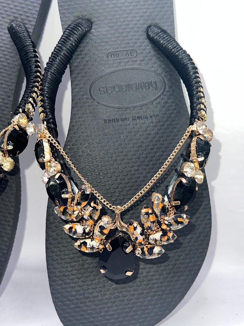 May include: Black flip-flops featuring black fabric-wrapped straps adorned with gold-tone chains and clusters of black and leopard-print faceted crystals. The footbed displays the brand name Havaianas and size 39-40, with a textured surface pattern.