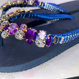 Puede incluir: Un par de chanclas azul marino con tiras envueltas en cinta de sat&eacute;n azul vibrante. Las tiras est&aacute;n adornadas con un grupo de cristales facetados morados y transparentes de varias formas, incluyendo cortes de pera y redondos, engastados en metal dorado.