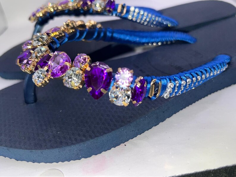 Puede incluir: Un par de chanclas azul marino con tiras envueltas en cinta de sat&eacute;n azul vibrante. Las tiras est&aacute;n adornadas con un grupo de cristales facetados morados y transparentes de varias formas, incluyendo cortes de pera y redondos, engastados en metal dorado.