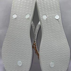 May include: A pair of white flip-flops with a textured sole and a thong strap. The straps are embellished with silver-toned rhinestones and a cluster of amber-colored crystals. The footbed features circular embossed details and size markings.