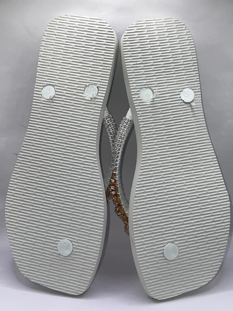 May include: A pair of white flip-flops with a textured sole and a thong strap. The straps are embellished with silver-toned rhinestones and a cluster of amber-colored crystals. The footbed features circular embossed details and size markings.