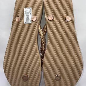 May include: A pair of tan flip-flops with a textured, ridged sole and a rose gold strap adorned with small, sparkling crystals. The left sandal features a white rectangular product label with a barcode and the text HAV SLIMFC.