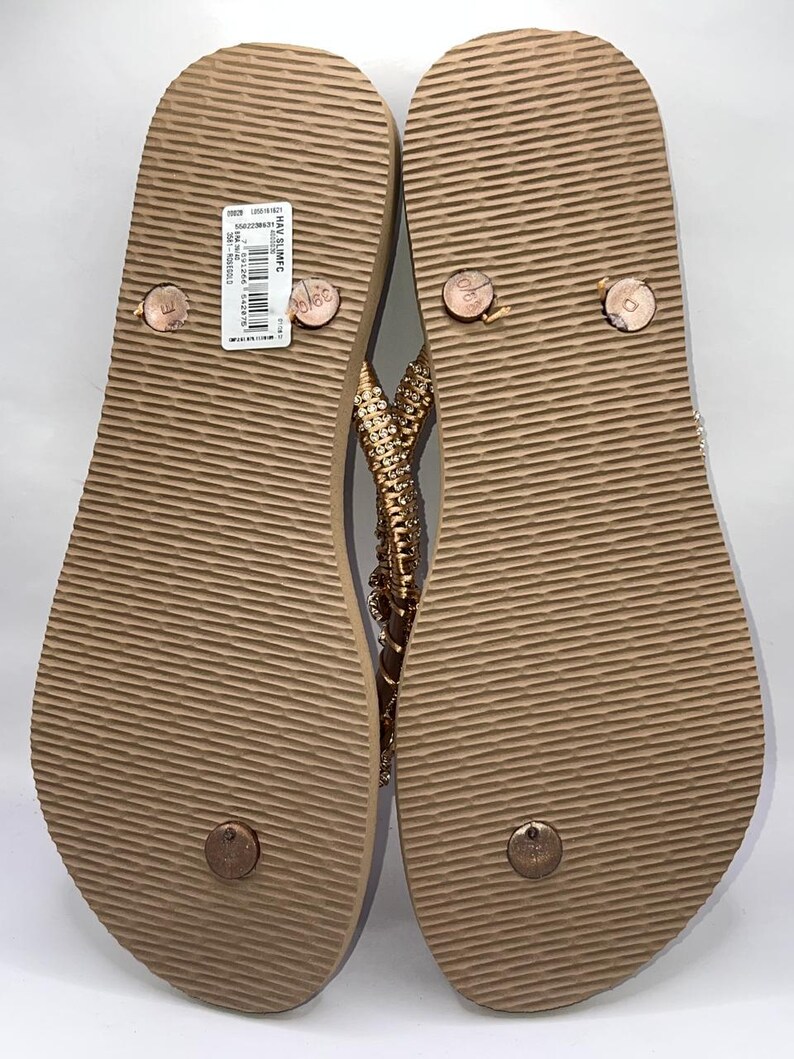 May include: A pair of tan flip-flops with a textured, ridged sole and a rose gold strap adorned with small, sparkling crystals. The left sandal features a white rectangular product label with a barcode and the text HAV SLIMFC.