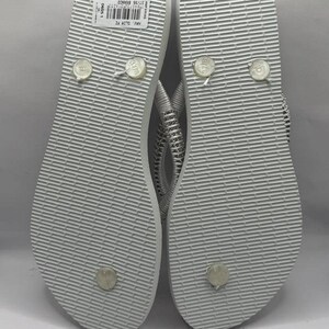 K&ouml;nnte beinhalten: Ein Paar wei&szlig;e Flip-Flops mit strukturierter Sohle. Die Riemen sind silber und wei&szlig;. Die Unterseite der Flip-Flops ist sichtbar und zeigt die Gr&ouml;&szlig;en- und Markeninformationen. Die Flip-Flops sind neu.