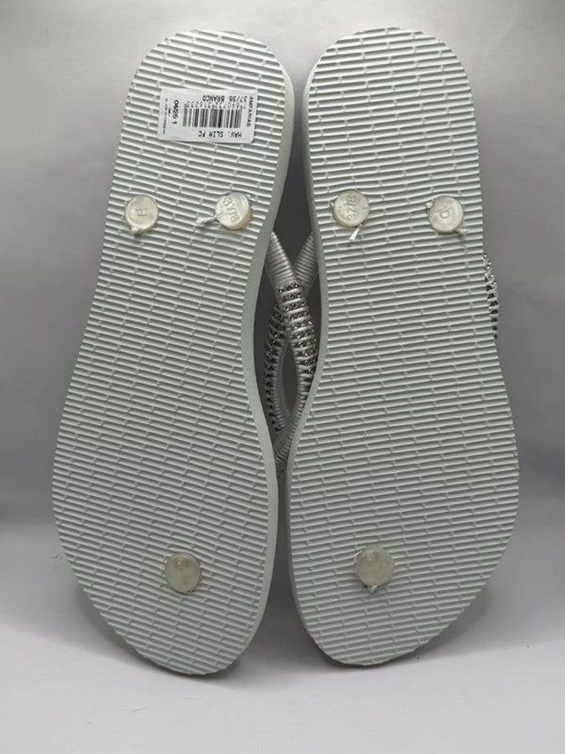 K&ouml;nnte beinhalten: Ein Paar wei&szlig;e Flip-Flops mit strukturierter Sohle. Die Riemen sind silber und wei&szlig;. Die Unterseite der Flip-Flops ist sichtbar und zeigt die Gr&ouml;&szlig;en- und Markeninformationen. Die Flip-Flops sind neu.