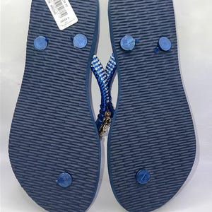 Puede incluir: Un par de chanclas azul marino con suela texturizada y una etiqueta blanca adherida al tal&oacute;n izquierdo. Las tiras est&aacute;n adornadas con filas de peque&ntilde;os cristales transparentes y dijes dorados, con un dise&ntilde;o delgado y tapones azules.