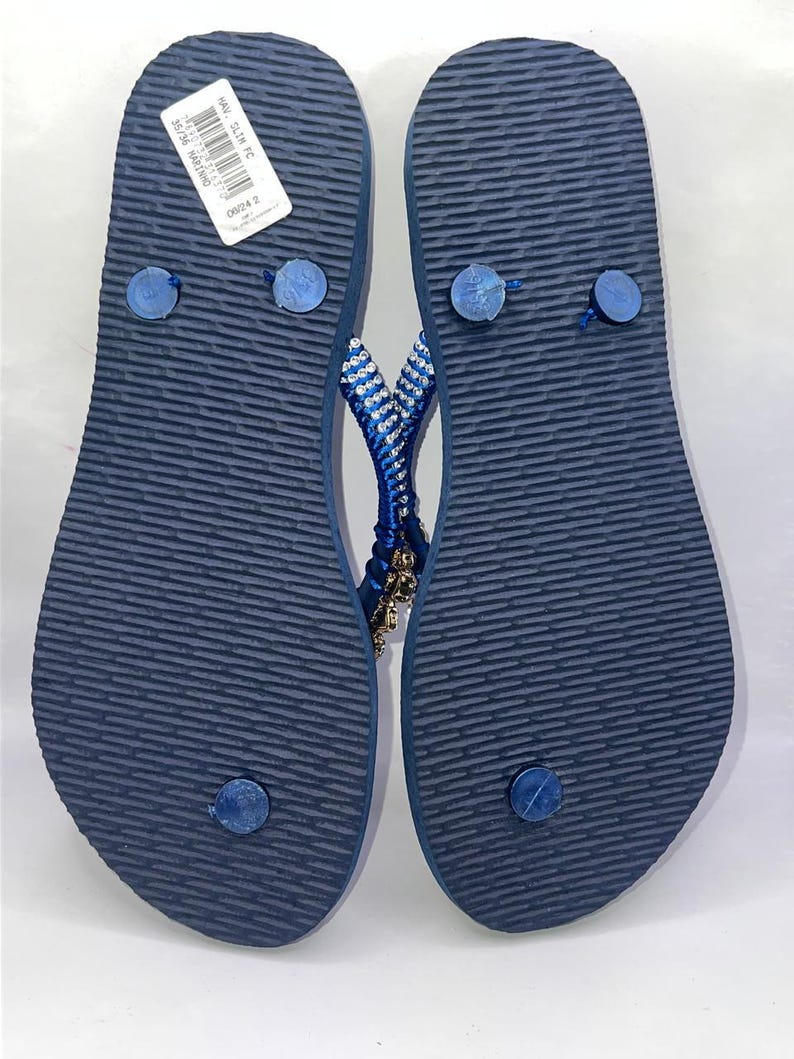 Puede incluir: Un par de chanclas azul marino con suela texturizada y una etiqueta blanca adherida al tal&oacute;n izquierdo. Las tiras est&aacute;n adornadas con filas de peque&ntilde;os cristales transparentes y dijes dorados, con un dise&ntilde;o delgado y tapones azules.