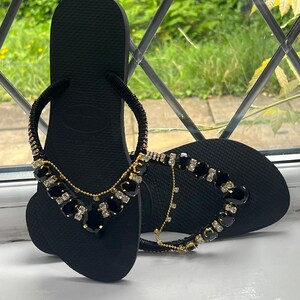 May include: Black flip-flops with decorative embellishments. The sandals feature a black base with a strap adorned with gold-toned chains and black and clear gemstones. The design adds a touch of elegance to the casual footwear.