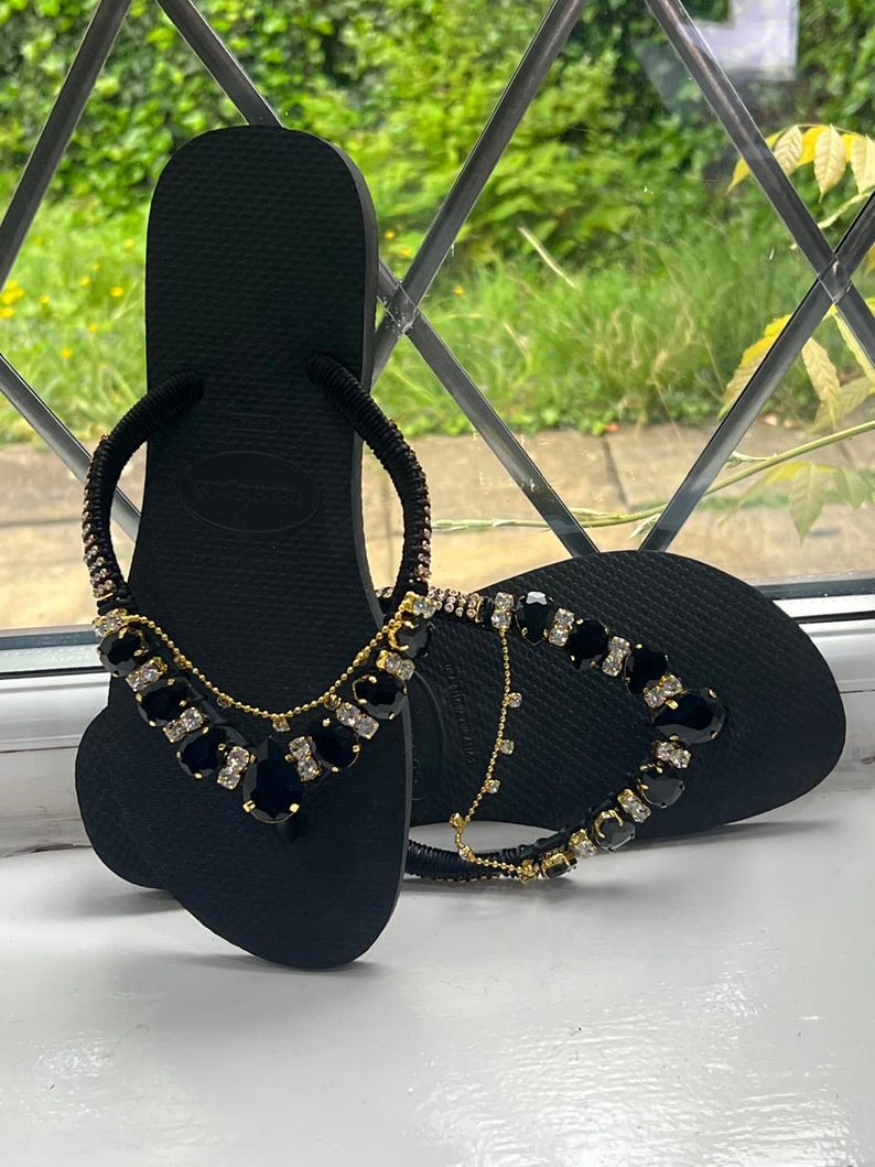 May include: Black flip-flops with decorative embellishments. The sandals feature a black base with a strap adorned with gold-toned chains and black and clear gemstones. The design adds a touch of elegance to the casual footwear.