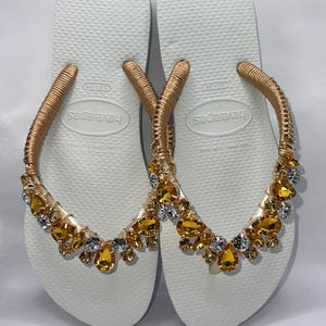 K&ouml;nnte beinhalten: Ein Paar wei&szlig;e Havaianas-Flip-Flops mit goldfarbenen Riemen, die mit goldenen und gelben Edelsteinen verziert sind. Die Flip-Flops sind in der Gr&ouml;&szlig;e 37-38 erh&auml;ltlich. Die Riemen sind mit goldfarbenem Material umwickelt.