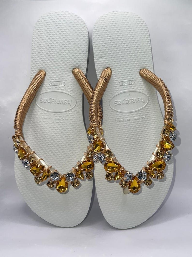 K&ouml;nnte beinhalten: Ein Paar wei&szlig;e Havaianas-Flip-Flops mit goldfarbenen Riemen, die mit goldenen und gelben Edelsteinen verziert sind. Die Flip-Flops sind in der Gr&ouml;&szlig;e 37-38 erh&auml;ltlich. Die Riemen sind mit goldfarbenem Material umwickelt.