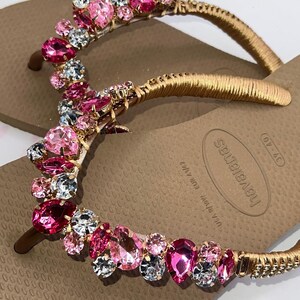 May include: A pair of tan flip-flops featuring gold-wrapped straps adorned with a cluster of faceted pink and clear crystals. The footbed displays the brand name Havaianas and the size 39-40, with USA 7/8W and EUR 41/42 printed on the surface.