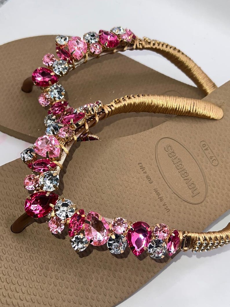 May include: A pair of tan flip-flops featuring gold-wrapped straps adorned with a cluster of faceted pink and clear crystals. The footbed displays the brand name Havaianas and the size 39-40, with USA 7/8W and EUR 41/42 printed on the surface.
