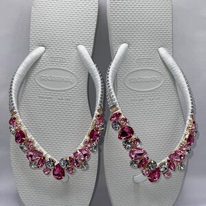 May include: A pair of white flip-flops featuring Havaianas branding on the footbed. The white straps are embellished with a collection of faceted pink and clear crystals in various shapes, securely attached to the top of each strap.