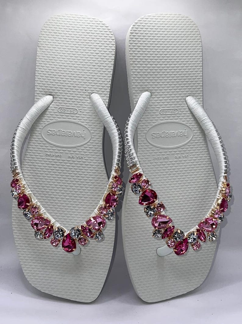 May include: A pair of white flip-flops featuring Havaianas branding on the footbed. The white straps are embellished with a collection of faceted pink and clear crystals in various shapes, securely attached to the top of each strap.