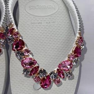 May include: A pair of white flip-flops featuring white fabric-wrapped straps adorned with a cluster of faceted pink and clear crystals in various shapes and sizes. The footbed displays the text Havaianas, USA 9/10, and EUR 41/42.