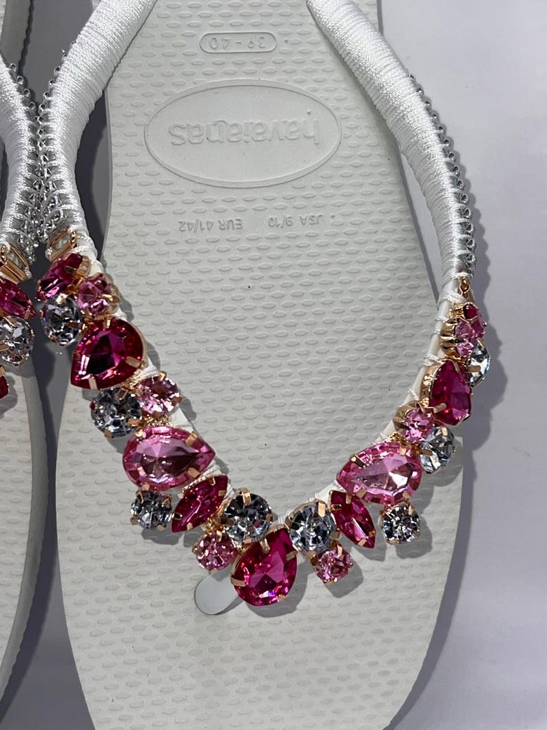 May include: A pair of white flip-flops featuring white fabric-wrapped straps adorned with a cluster of faceted pink and clear crystals in various shapes and sizes. The footbed displays the text Havaianas, USA 9/10, and EUR 41/42.