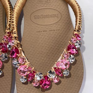 May include: A pair of beige flip-flops featuring gold-wrapped straps adorned with an array of faceted pink and clear crystals. The footbed displays the brand name Havaianas and size markings 39-40, USA 9/10W, and EUR 41/42 in a textured rubber finish.