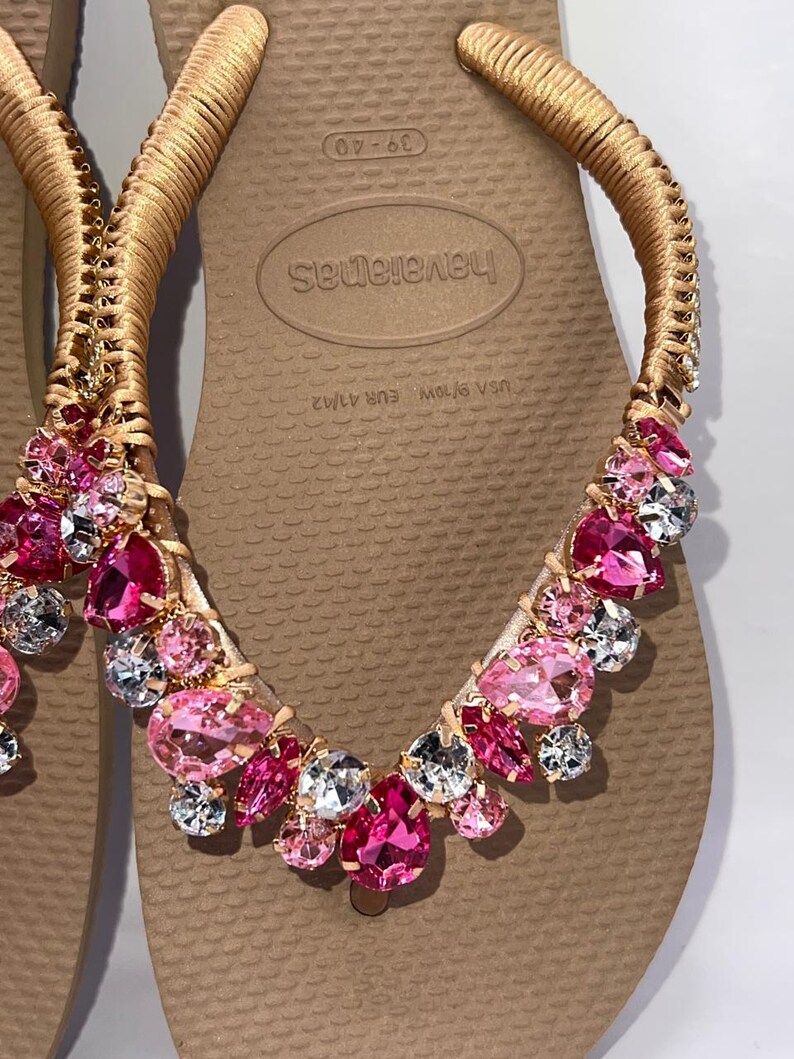 May include: A pair of beige flip-flops featuring gold-wrapped straps adorned with an array of faceted pink and clear crystals. The footbed displays the brand name Havaianas and size markings 39-40, USA 9/10W, and EUR 41/42 in a textured rubber finish.