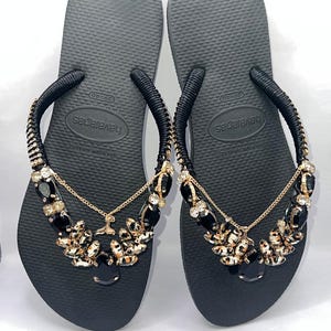 May include: A pair of black flip-flops featuring black fabric-wrapped straps adorned with gold-tone chains and clusters of black and clear faceted crystals. The footbeds are textured with a rice-grain pattern and display the Havaianas brand logo.