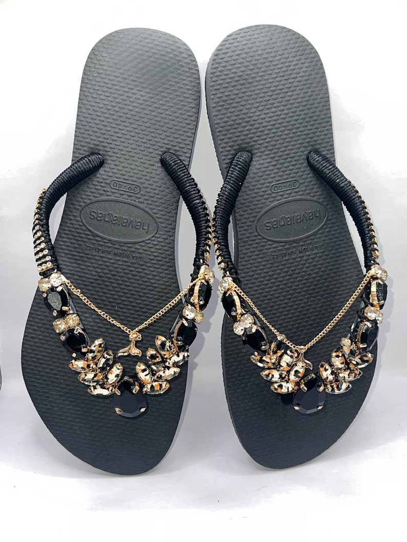 May include: A pair of black flip-flops featuring black fabric-wrapped straps adorned with gold-tone chains and clusters of black and clear faceted crystals. The footbeds are textured with a rice-grain pattern and display the Havaianas brand logo.