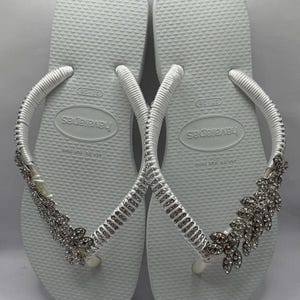 K&ouml;nnte beinhalten: Wei&szlig;e Havaianas-Flip-Flops mit silbernen Strassverzierungen. Die Riemen sind wei&szlig; und mit Strasssteinen verziert. Die Flip-Flops haben ein Blumendesign an der Seite.