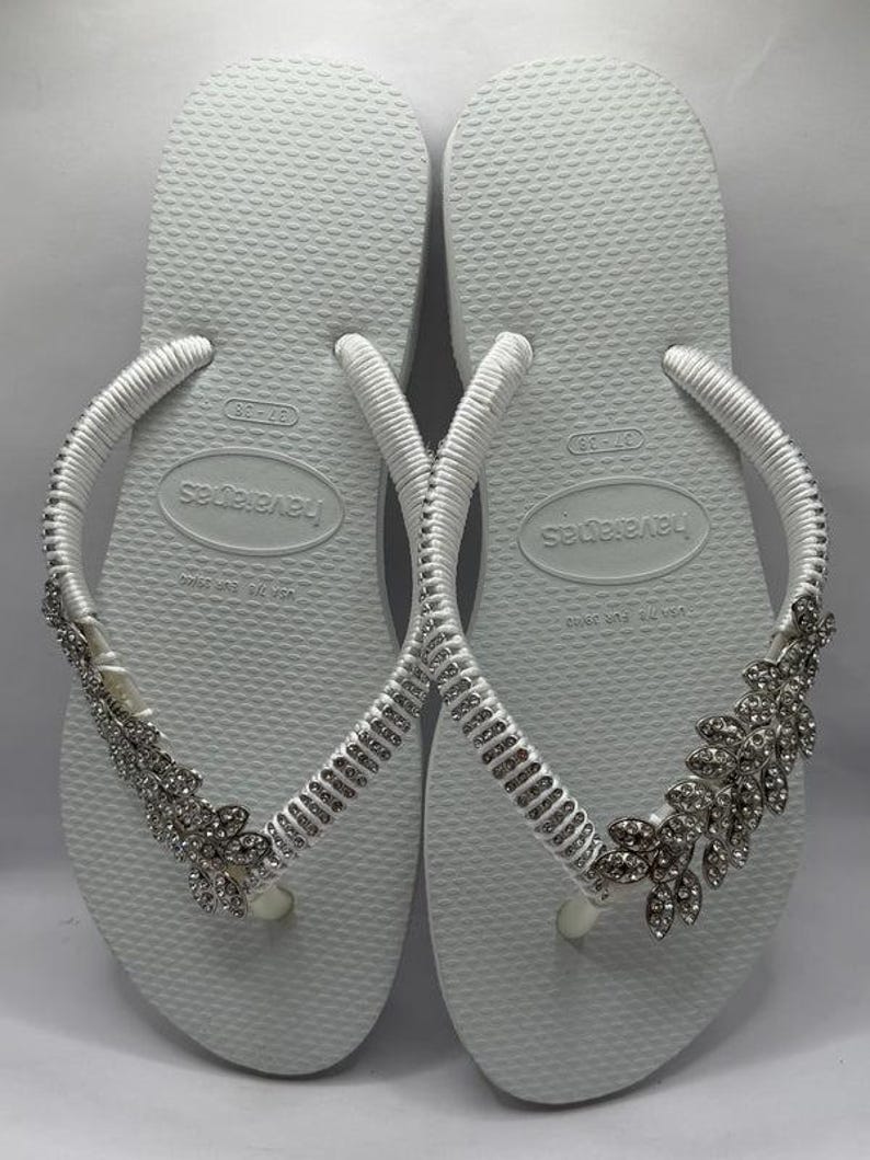 K&ouml;nnte beinhalten: Wei&szlig;e Havaianas-Flip-Flops mit silbernen Strassverzierungen. Die Riemen sind wei&szlig; und mit Strasssteinen verziert. Die Flip-Flops haben ein Blumendesign an der Seite.