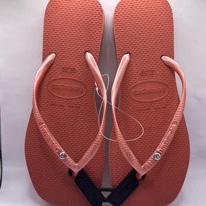 K&ouml;nnte beinhalten: Paar korallenfarbene Havaianas-Flip-Flops. Die Sandalen haben ein strukturiertes Fu&szlig;bett, passende Riemen und den Markennamen auf der Seite aufgedruckt. Ein schwarzes Etikett ist zwischen den Sandalen befestigt.