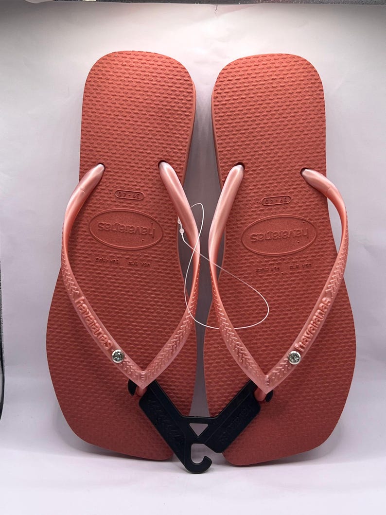 K&ouml;nnte beinhalten: Paar korallenfarbene Havaianas-Flip-Flops. Die Sandalen haben ein strukturiertes Fu&szlig;bett, passende Riemen und den Markennamen auf der Seite aufgedruckt. Ein schwarzes Etikett ist zwischen den Sandalen befestigt.