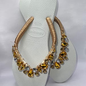 K&ouml;nnte beinhalten: Wei&szlig;e Havaianas-Flip-Flops mit goldenen Riemen, die mit bernsteinfarbenen und klaren Edelsteinen verziert sind. Die Sandalen haben eine strukturierte Sohle und sind f&uuml;r den l&auml;ssigen Gebrauch konzipiert. Die Edelsteine verleihen dem schlichten Design einen Hauch von Eleganz.