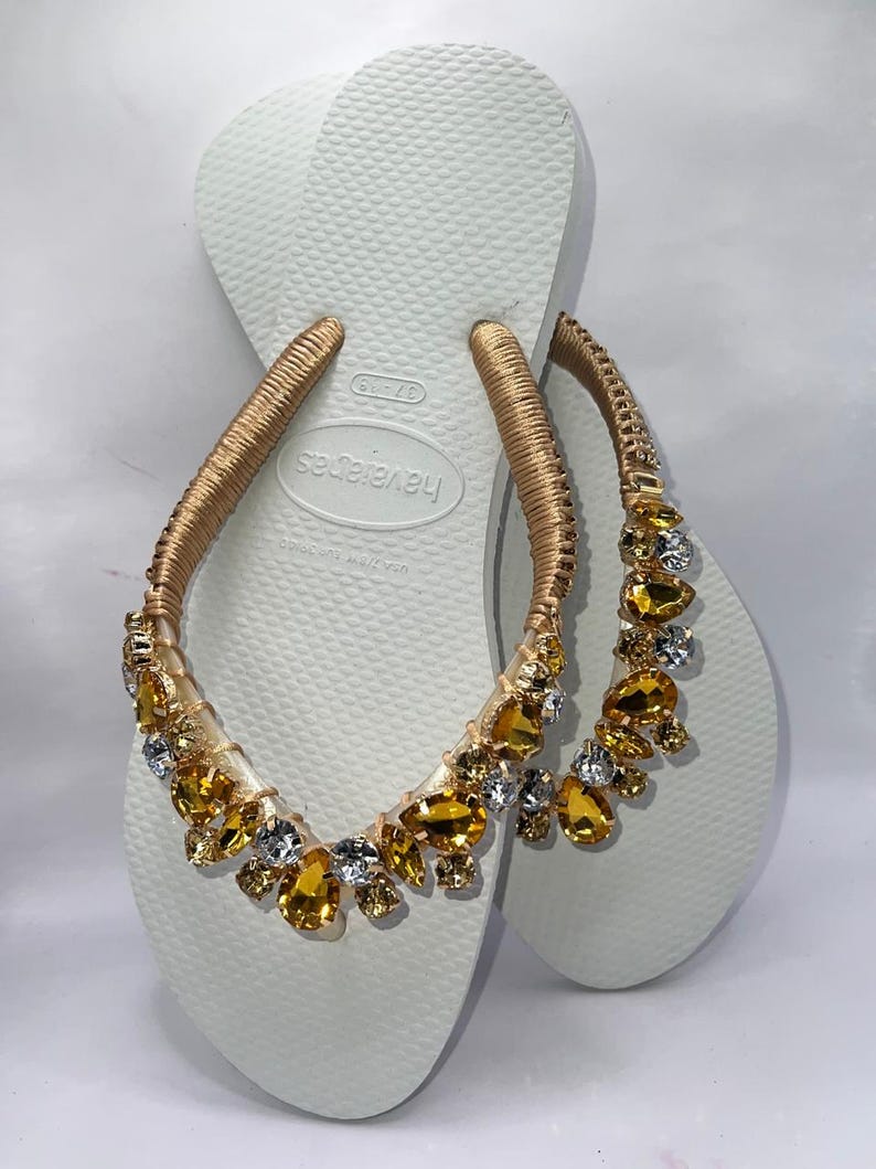K&ouml;nnte beinhalten: Wei&szlig;e Havaianas-Flip-Flops mit goldenen Riemen, die mit bernsteinfarbenen und klaren Edelsteinen verziert sind. Die Sandalen haben eine strukturierte Sohle und sind f&uuml;r den l&auml;ssigen Gebrauch konzipiert. Die Edelsteine verleihen dem schlichten Design einen Hauch von Eleganz.