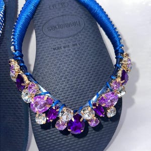 Puede incluir: Un par de chanclas azul marino con tiras envueltas en tela azul brillante. Las tiras est&aacute;n adornadas con un grupo de cristales facetados de color p&uacute;rpura, violeta y transparente engastados en metal dorado. La plantilla muestra el texto Havaianas USA 6W EUR 37/38.