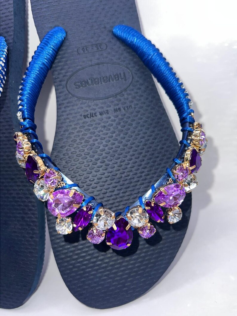 Puede incluir: Un par de chanclas azul marino con tiras envueltas en tela azul brillante. Las tiras est&aacute;n adornadas con un grupo de cristales facetados de color p&uacute;rpura, violeta y transparente engastados en metal dorado. La plantilla muestra el texto Havaianas USA 6W EUR 37/38.
