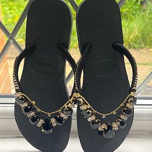 May include: Black Havaianas flip-flops with embellished straps. The straps are adorned with black and clear gemstones and gold-colored chain. The flip-flops are displayed on a white surface with a window in the background.