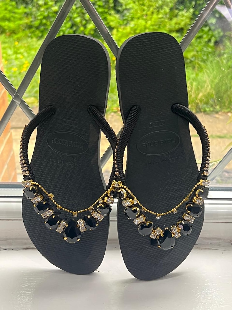 May include: Black Havaianas flip-flops with embellished straps. The straps are adorned with black and clear gemstones and gold-colored chain. The flip-flops are displayed on a white surface with a window in the background.