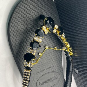 May include: Black Havaianas flip-flops with a decorative strap. The strap is embellished with black and clear gemstones, and gold-colored accents. The flip-flops are size 39-40.