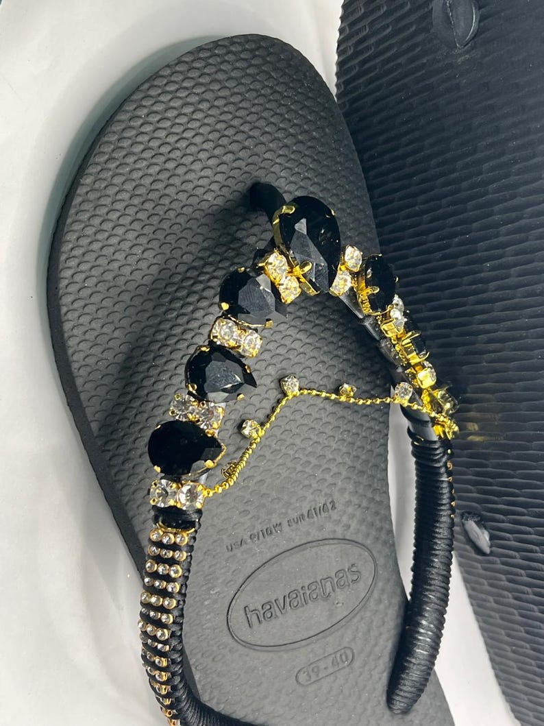 May include: Black Havaianas flip-flops with a decorative strap. The strap is embellished with black and clear gemstones, and gold-colored accents. The flip-flops are size 39-40.