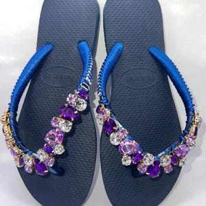 Puede incluir: Un par de chanclas azul marino con tiras envueltas en tela azul, adornadas con un grupo de cristales facetados de color morado y transparente. Las plantillas tienen una textura de grano de arroz y muestran la marca Havaianas en el centro.