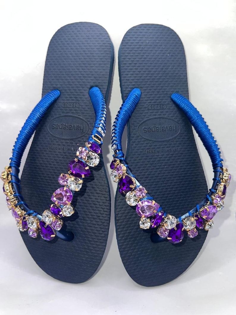 Puede incluir: Un par de chanclas azul marino con tiras envueltas en tela azul, adornadas con un grupo de cristales facetados de color morado y transparente. Las plantillas tienen una textura de grano de arroz y muestran la marca Havaianas en el centro.