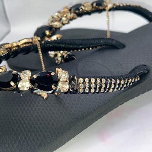 May include: A black flip-flop sandal featuring a black strap adorned with teardrop-shaped black crystals, round champagne-coloured rhinestones, and rows of small clear crystals. The strap is wrapped in black thread, and a thin gold chain is attached.