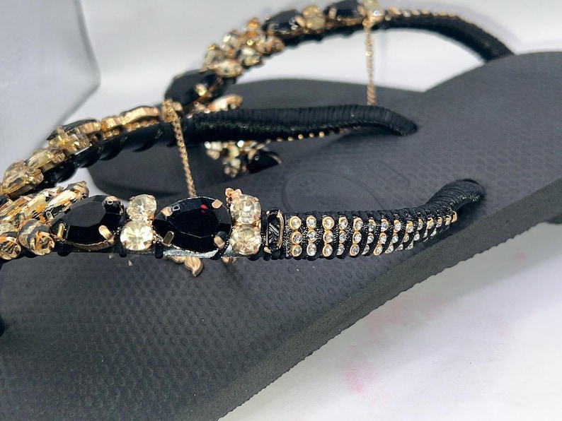 May include: A black flip-flop sandal featuring a black strap adorned with teardrop-shaped black crystals, round champagne-coloured rhinestones, and rows of small clear crystals. The strap is wrapped in black thread, and a thin gold chain is attached.