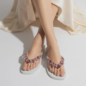 May include: A pair of white flip-flops featuring straps adorned with a cluster of faceted pink and clear crystals. The footwear has a flat sole and a thong-style design, with the decorative embellishments covering the top of the straps.