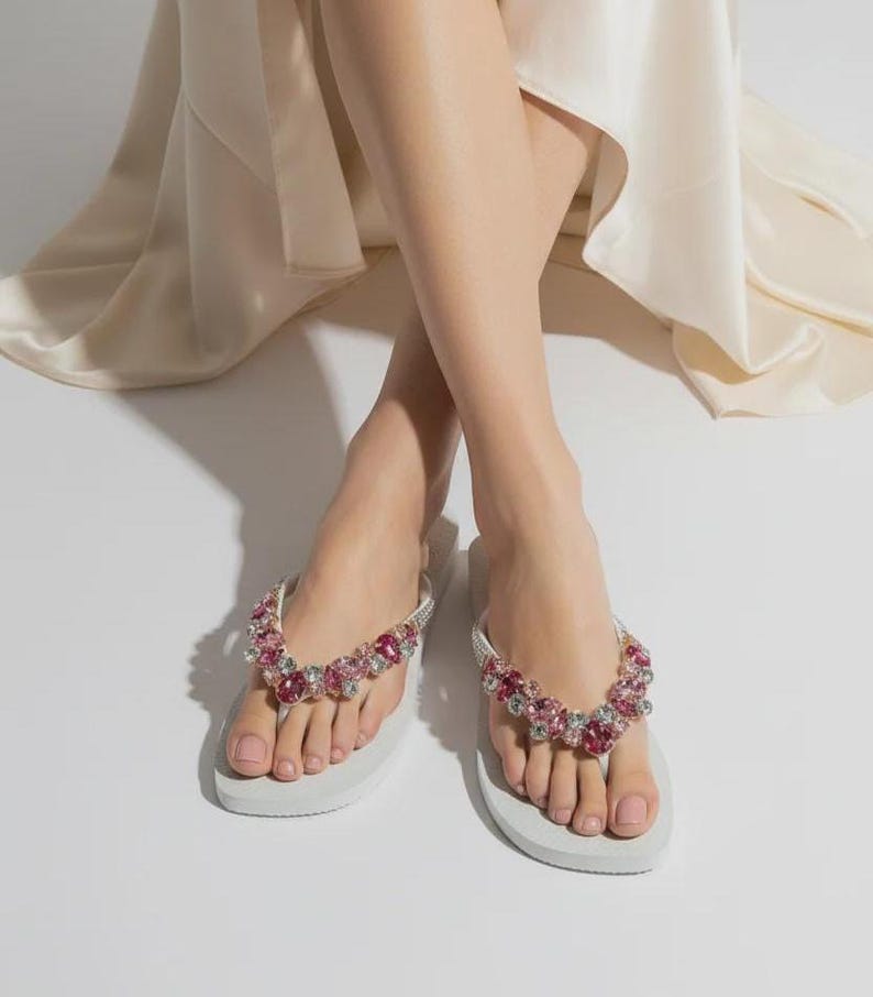 May include: A pair of white flip-flops featuring straps adorned with a cluster of faceted pink and clear crystals. The footwear has a flat sole and a thong-style design, with the decorative embellishments covering the top of the straps.