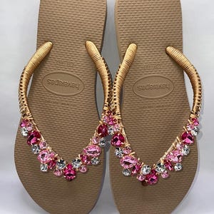 May include: A pair of tan flip-flops featuring gold-wrapped straps adorned with a cluster of faceted pink and clear crystals. The footbeds are textured and display the Havaianas brand name along with the size 39-40 printed in the center.