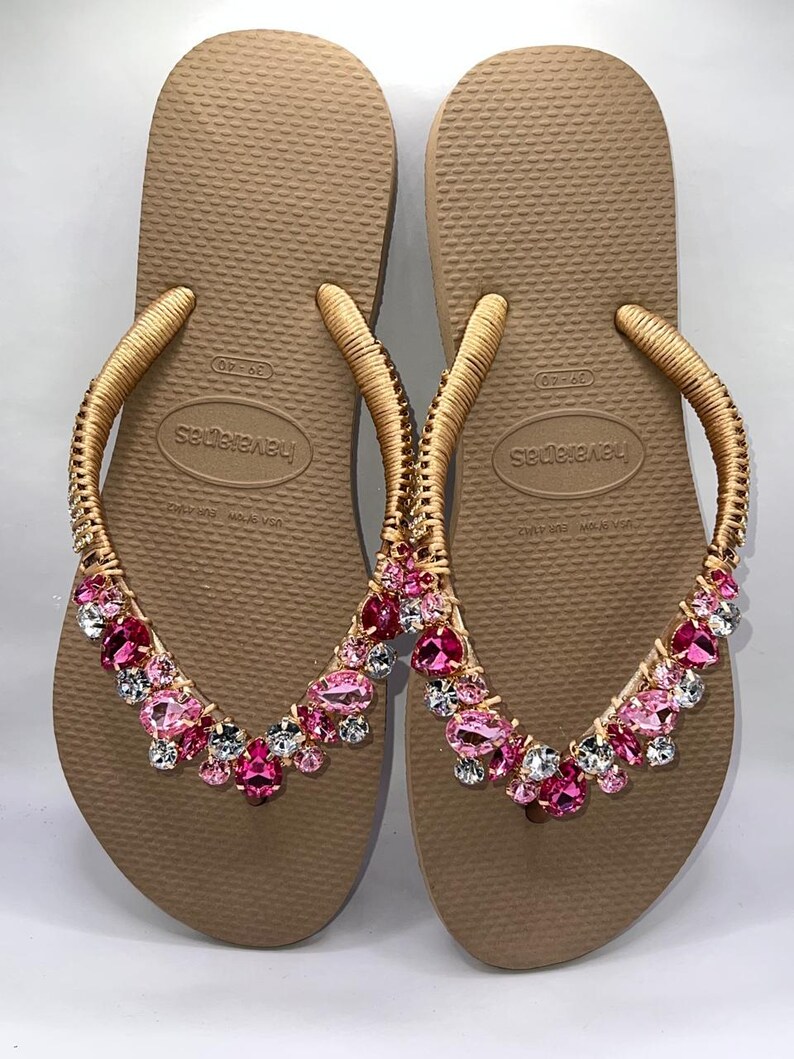 May include: A pair of tan flip-flops featuring gold-wrapped straps adorned with a cluster of faceted pink and clear crystals. The footbeds are textured and display the Havaianas brand name along with the size 39-40 printed in the center.