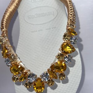 K&ouml;nnte beinhalten: Wei&szlig;e Havaianas-Zehensandale mit goldfarbenem Riemen, verziert mit goldenen, gelben und klaren Edelsteinen. Der Riemen ist mit goldfarbenem Material umwickelt. Die Zehensandale ist Gr&ouml;&szlig;e 37-38, was der EU-Gr&ouml;&szlig;e 39/40 entspricht.